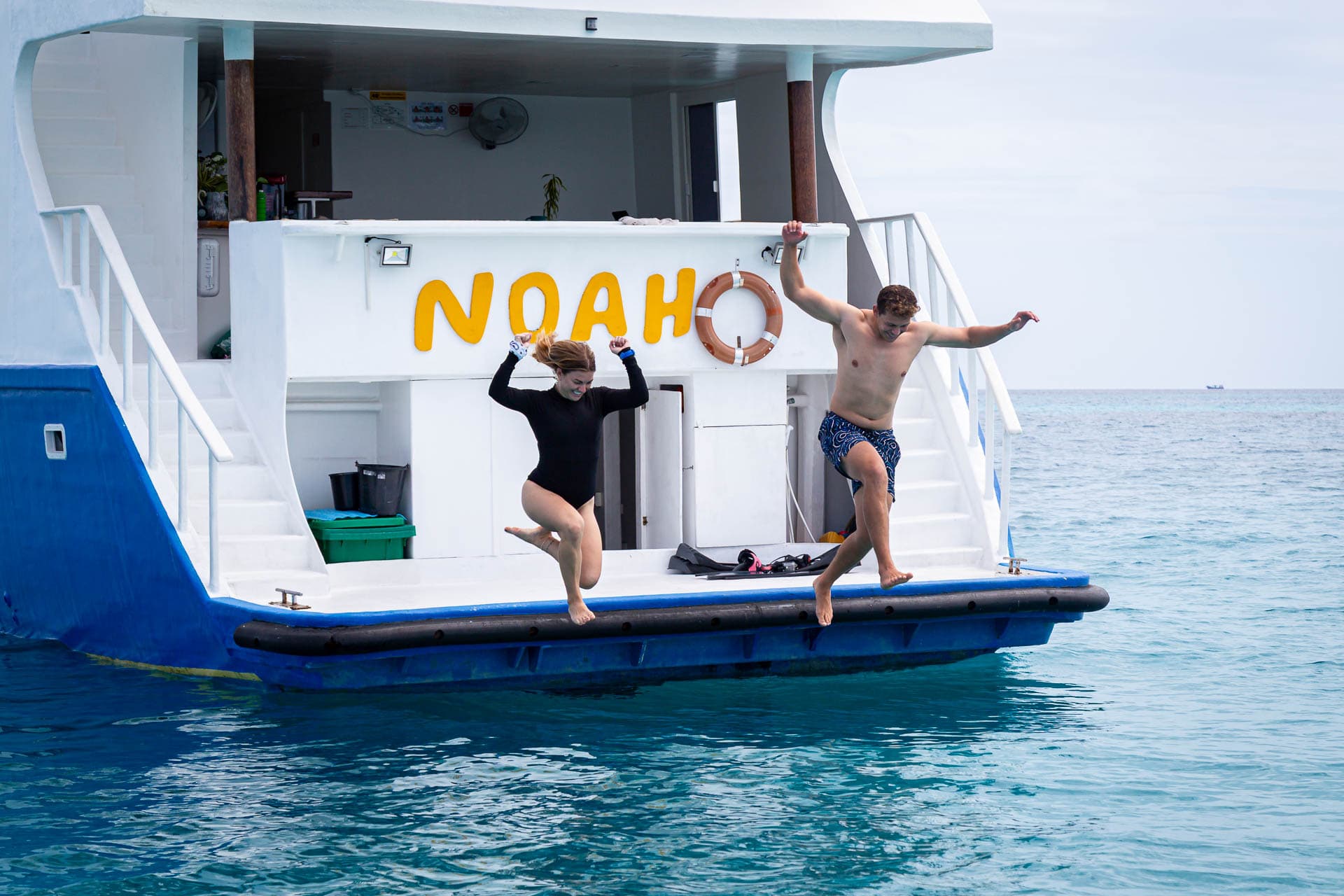 Two guests jumping off the swim platform under the yellow NOAH lettering on the stern